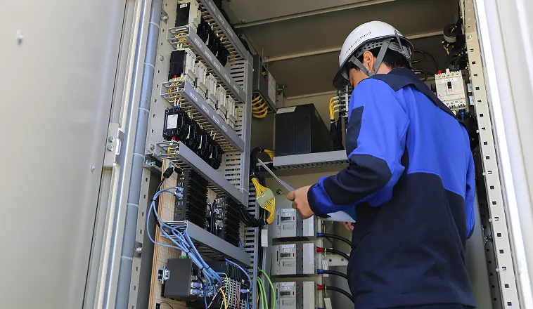 制御盤のある設備内で、ヘルメットと作業服を着た作業員が機器の点検・メンテナンスをしている様子。