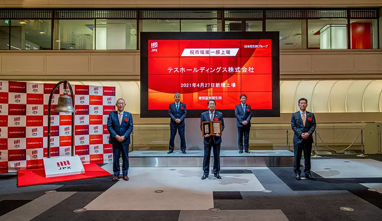 JPX（日本取引所グループ）の会場で、上場の記念式典として関係者が壇上に並び、大型スクリーンに社名が表示されている様子。