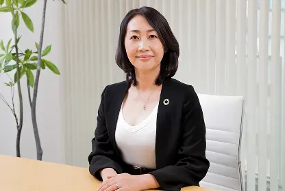 黒いジャケットと白いトップスを着用し、左胸にカラフルな円形のピンバッジを付けた女性が、木の机に手を組んで微笑みながら正面を向いているポートレート。背景に観葉植物とブラインドの窓。