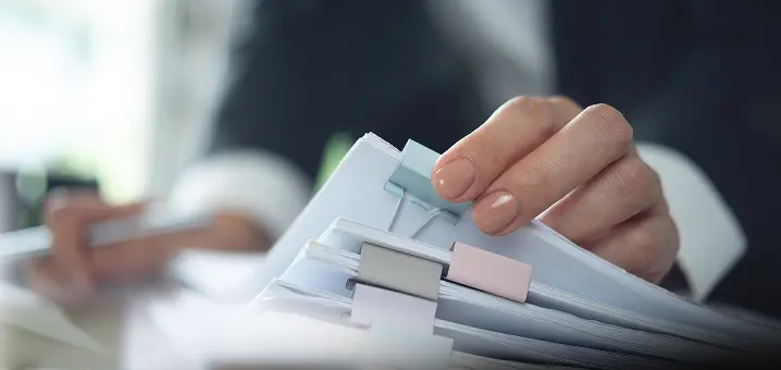 パステルカラーのクリップで束ねられた白い書類の山に、手が触れているクローズアップ。背景はぼかしたオフィス。