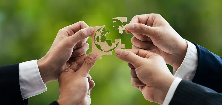 スーツ姿の4人が、緑色の世界地図が描かれた4枚のジグソーパズルを組み立てている様子。背景はぼかした緑の木々。
