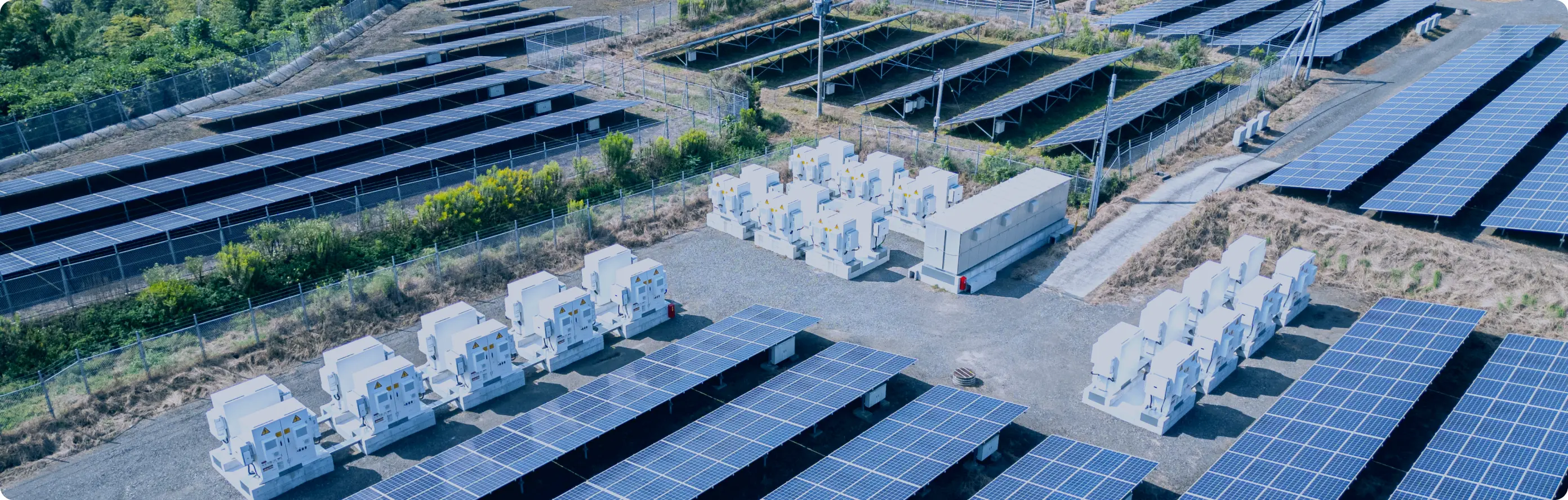 整列したパネルのあいだに白いコンテナ型の蓄電・電力設備が配置されている空からの風景
