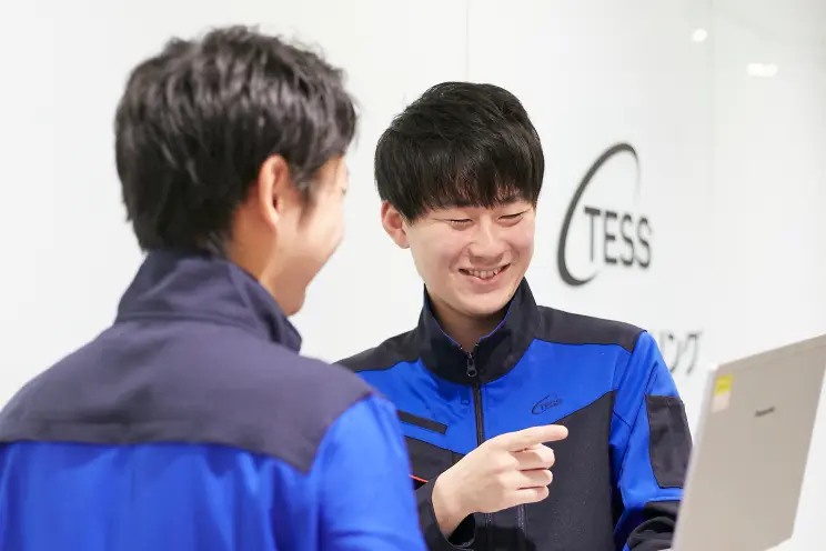 作業服を着た2人の社員が、壁のTESSロゴの前でノートパソコンを囲み、画面を指さしながら打ち合わせしている様子。