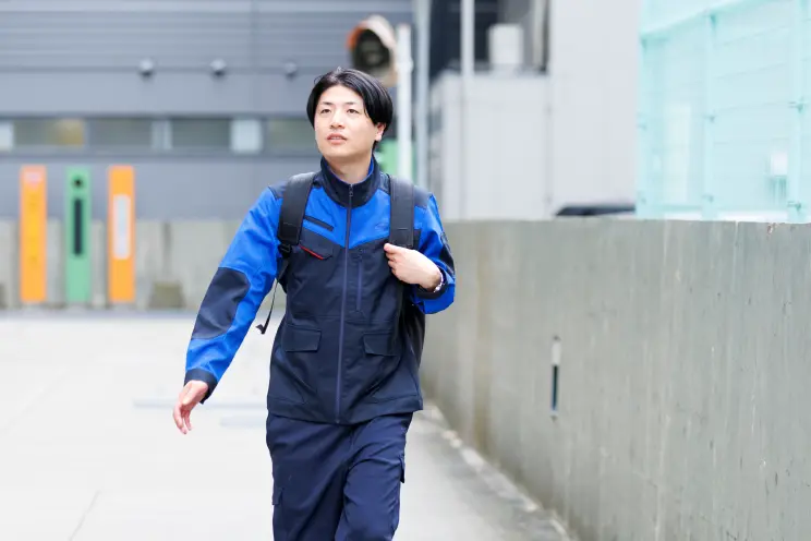 青と紺の作業服にリュックを背負った社員が、屋外の工場や倉庫街のような通路を歩いている様子。