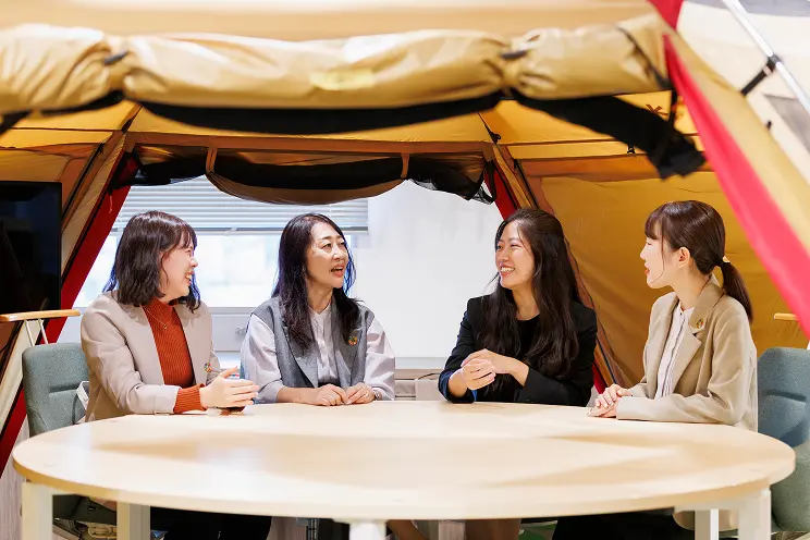 ベージュのテント内で、円卓を囲む4人の女性が談笑しながら打ち合わせをしている様子。