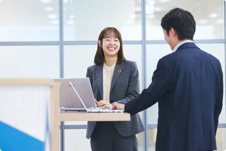 スタンディングデスクの両側に立つ男女が、ノートパソコンを開いて話している様子。女性のスーツにはSDGsを思わせる円形のバッジが付いている。