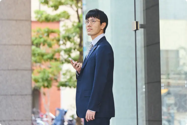 ネイビーのスーツ姿の男性が、屋外のビジネス街でスマートフォンを持ち、視線を上に向けている様子。
