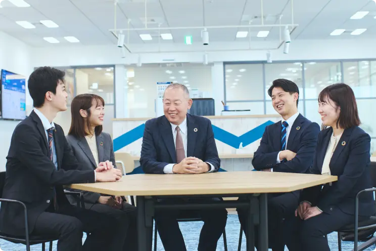 明るいオフィスの会議室で、五角形のテーブルを囲む5人のビジネスパーソンが笑顔で話し合っている様子。全員がスーツにSDGsを思わせる円形のピンバッジを付けている。